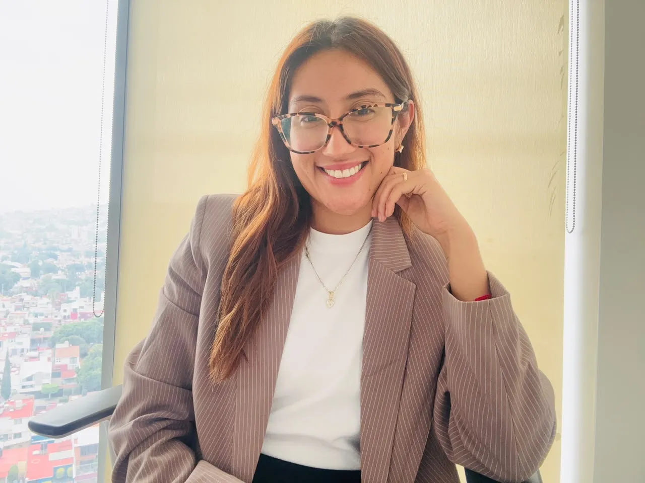Geraldine Ramírez sentada junto a una ventana con vista a la ciudad, con saco de rayas y blusa blanca.